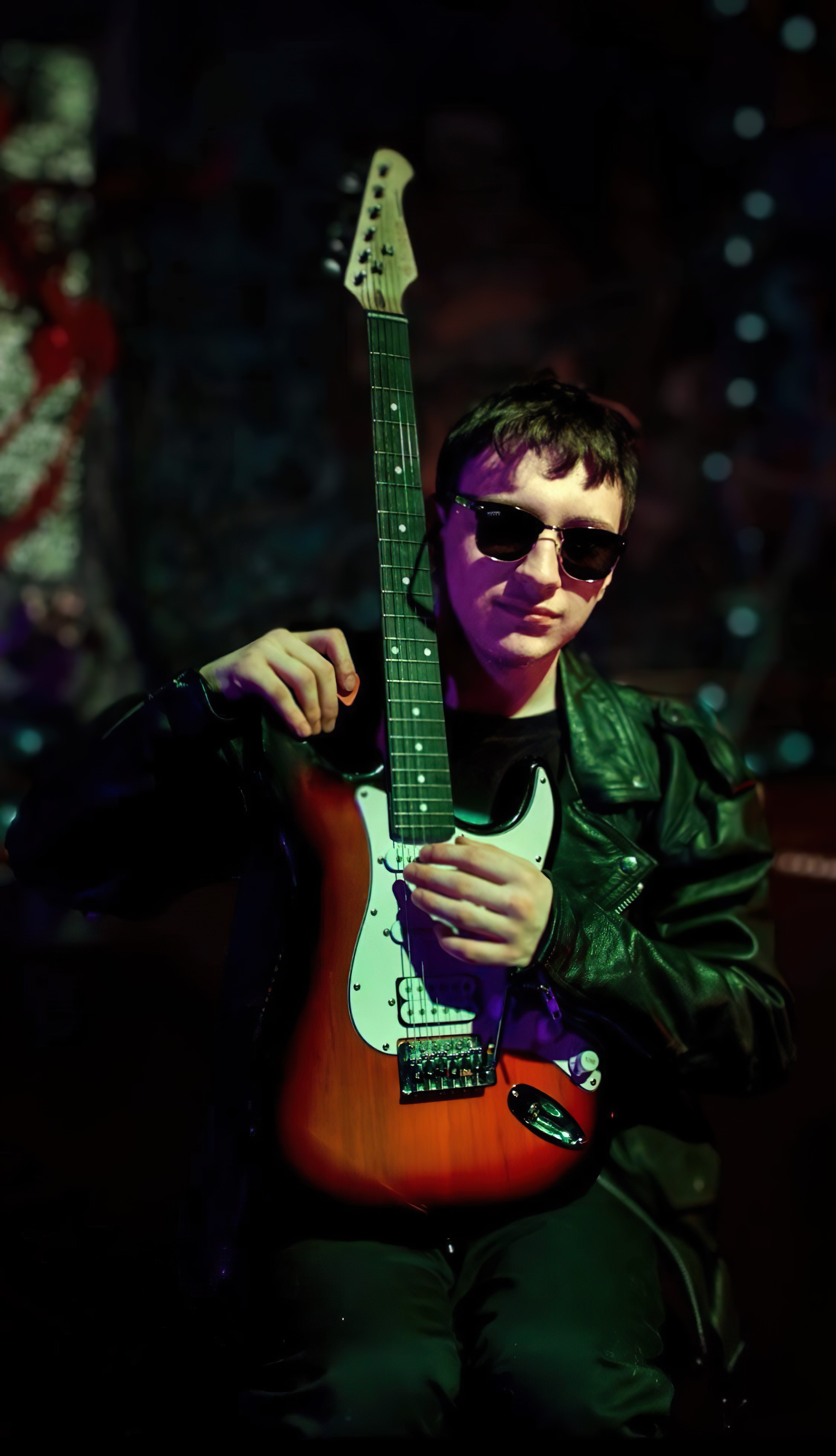A young musician in sunglasses holding an electric guitar in a moody, dark atmosphere.