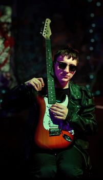 A young musician in sunglasses holding an electric guitar in a moody, dark atmosphere.