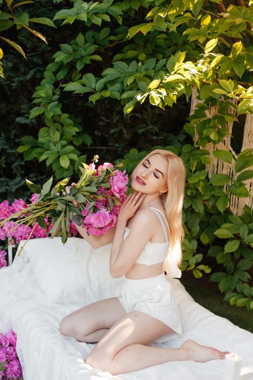 Free Beautiful blonde woman posing with lush pink peonies in a serene outdoor setting. Ideal for fashion and lifestyle themes. Stock Photo