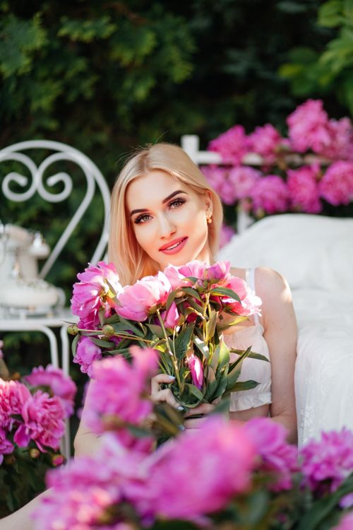Free Elegant blonde woman smiling while holding pink peonies in a garden, surrounded by floral beauty. Stock Photo