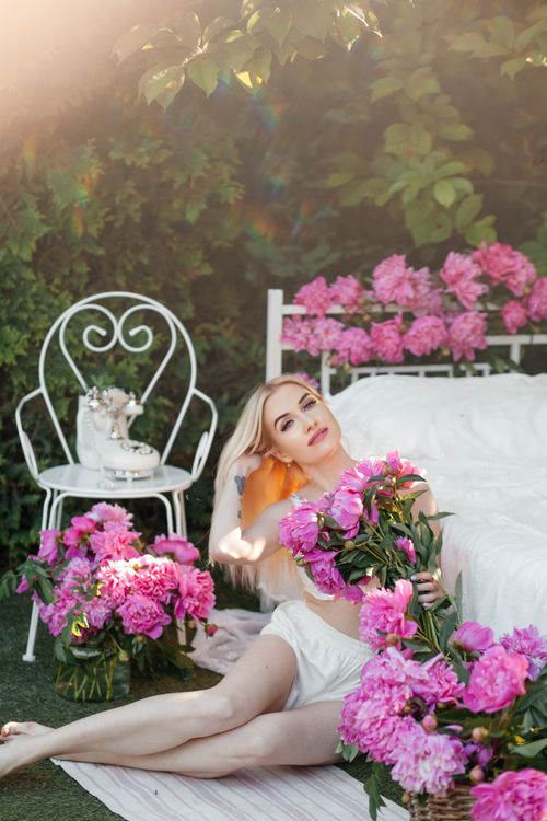 Free Blonde woman in white pajamas posing with pink flowers in a sunlit garden. Stock Photo