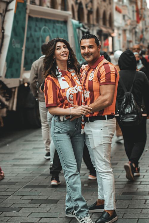 Free A happy couple wearing sports jerseys poses on a lively city street, showcasing team spirit and vibrant fashion. Stock Photo