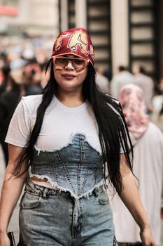 Young woman in denim outfit and cap walking through busy city street.