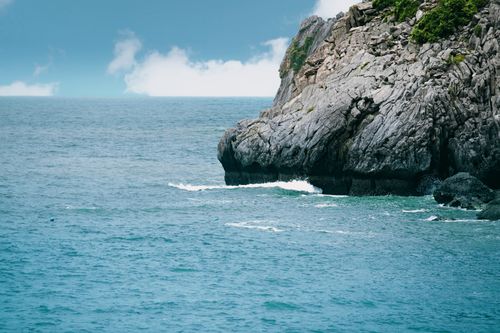 Free A striking rocky sea cliff with blue ocean waves and cloud-dappled sky. Stock Photo