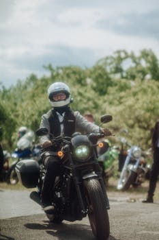 Man riding a motorcycle with a white helmet in a sunny outdoor setting with several motorbikes in the background.