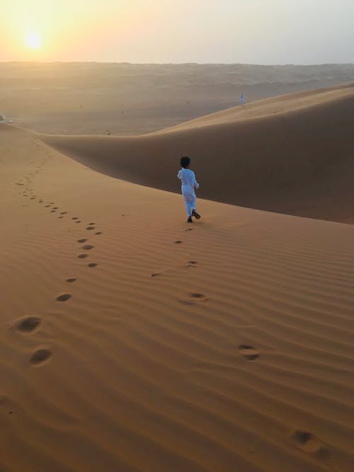ゴールデンアワーの間に砂漠の砂の上を一人で歩いている白いトーブのイスラム教徒の少年の背面図の写真