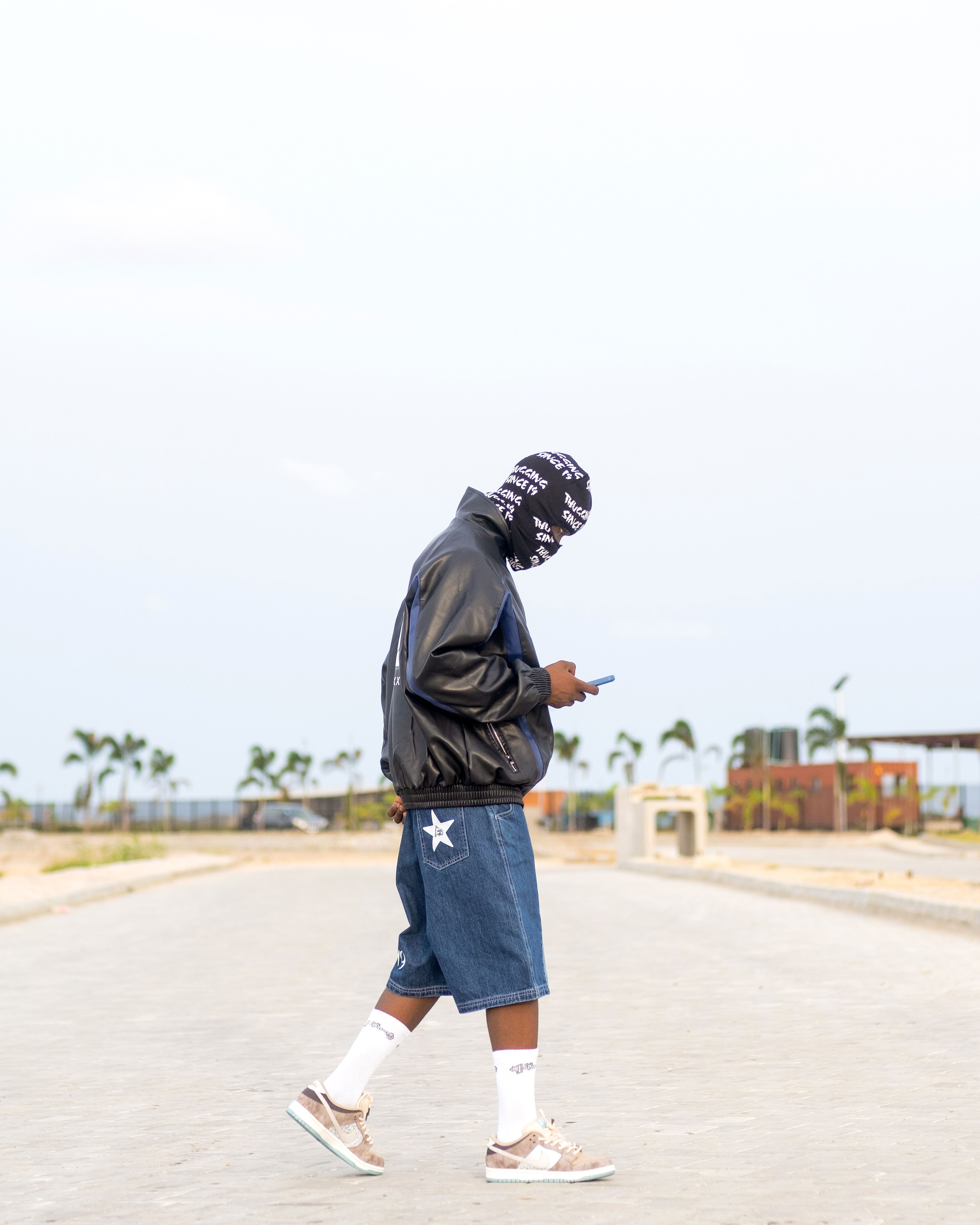 Stylish man in a balaclava and denim shorts walking on an empty street checking his phone.