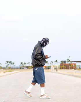 Stylish man in a balaclava and denim shorts walking on an empty street checking his phone.