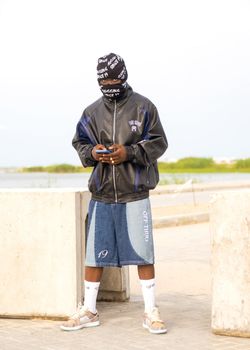 A man in street style fashion with a mask, jacket, and shorts holds a smartphone outdoors.