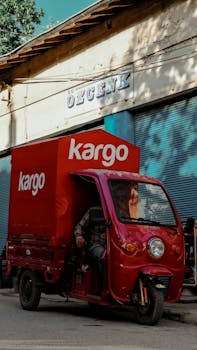A red auto rickshaw labeled 'Kargo' parked outside an urban building under sunlight.