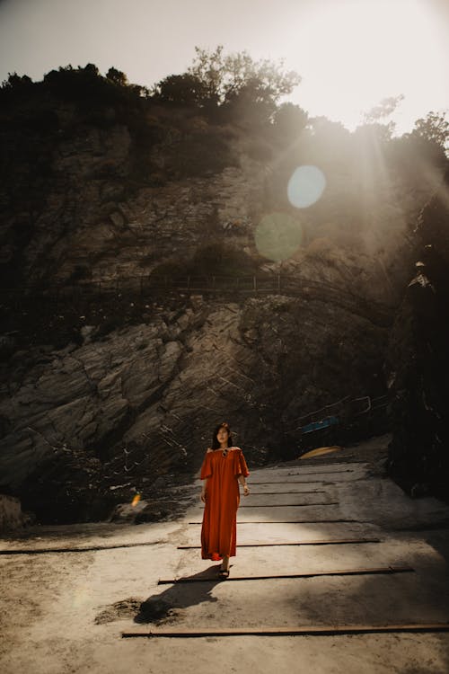 Woman in Orange Dress Standing on Pathway Below A Rock Formation