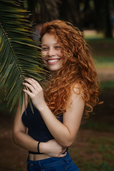 Portrait of a happy woman with curly hair smiling while holding a palm leaf outdoors.