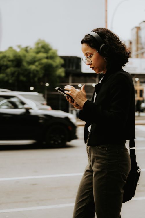 Free Woman walking in city street, wearing headphones and using smartphone, showcasing modern urban lifestyle. Stock Photo