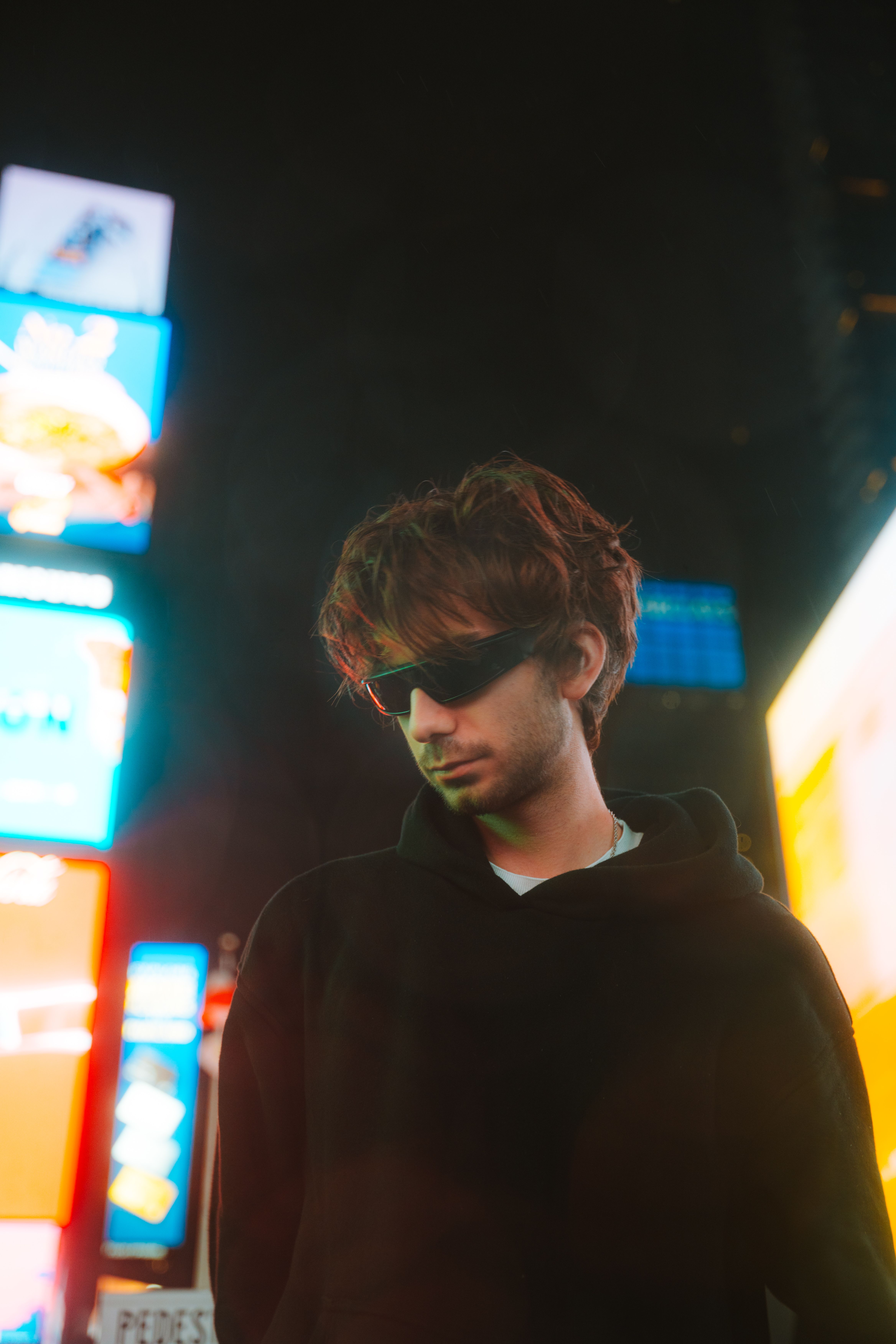 Portrait of a fashionable man in a black hoodie and sunglasses, captured at night in a vibrant urban setting.