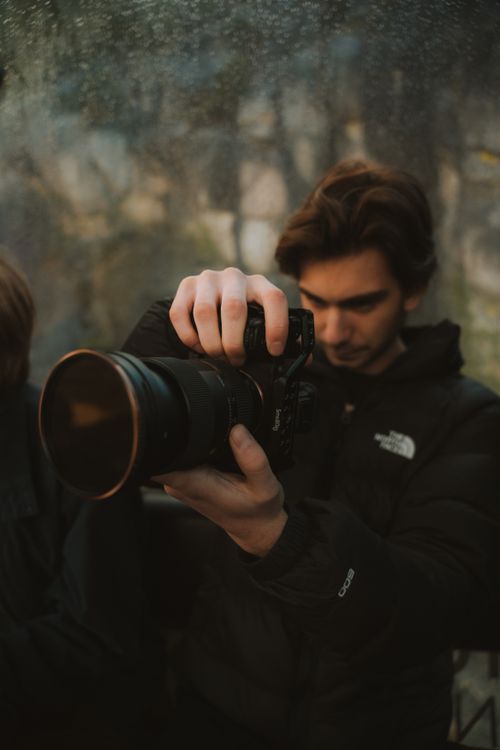 Free A young adult in a black jacket holds a digital camera outdoors, focusing intently. Stock Photo