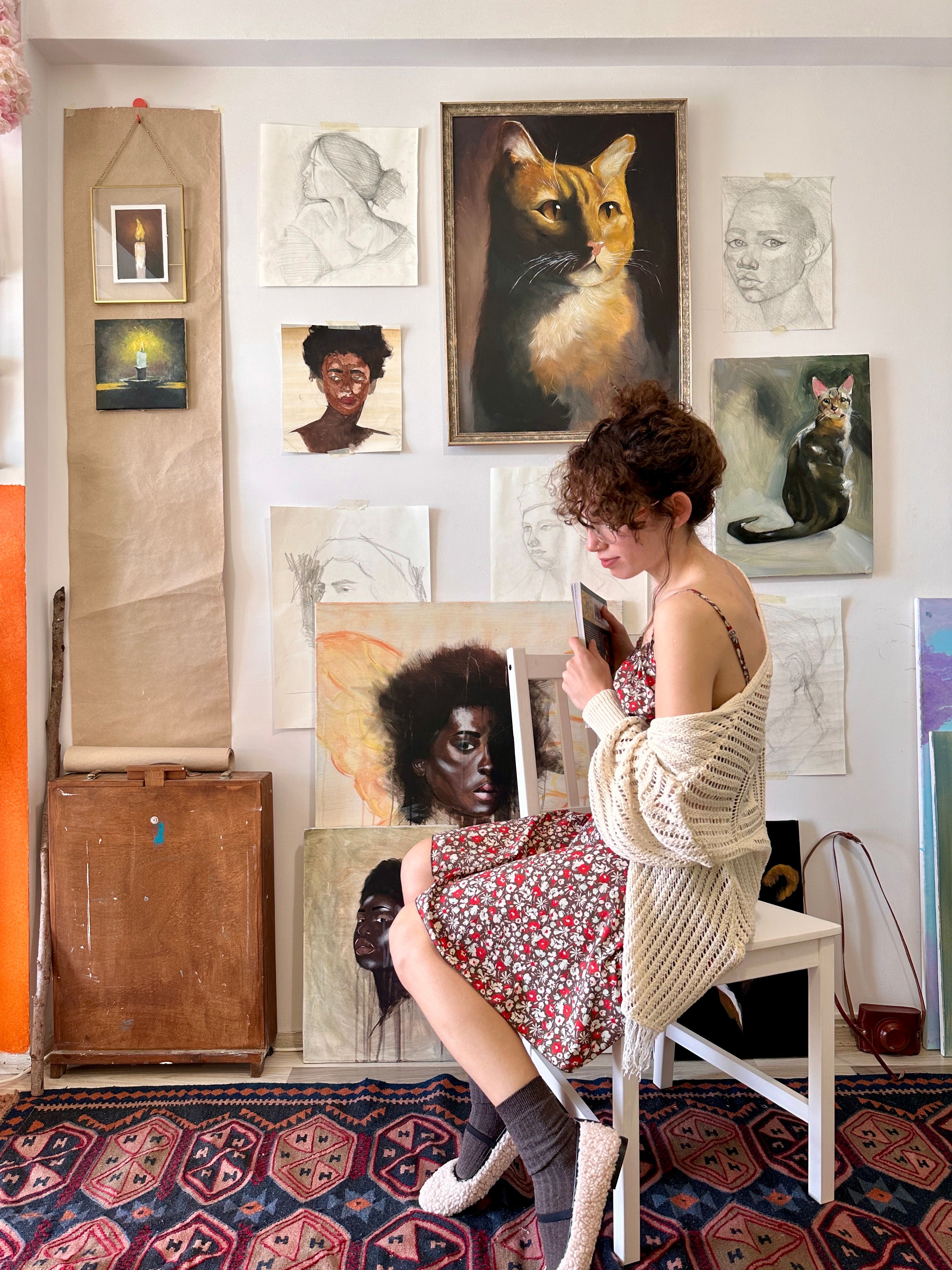 A woman sitting in an art studio surrounded by diverse portraits and artwork.