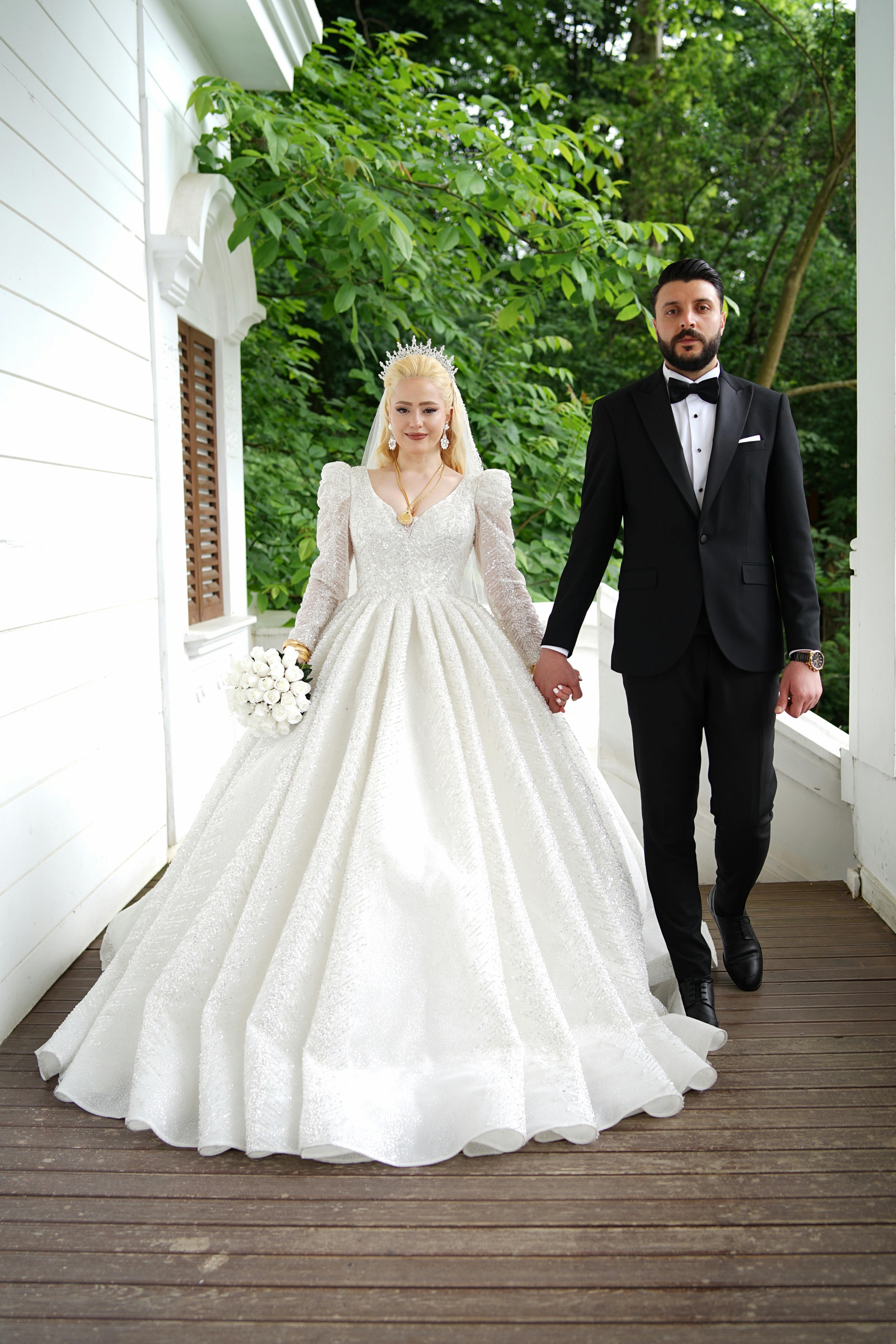 A bride and groom in formal attire holding hands on a wooden walkway surrounded by greenery.