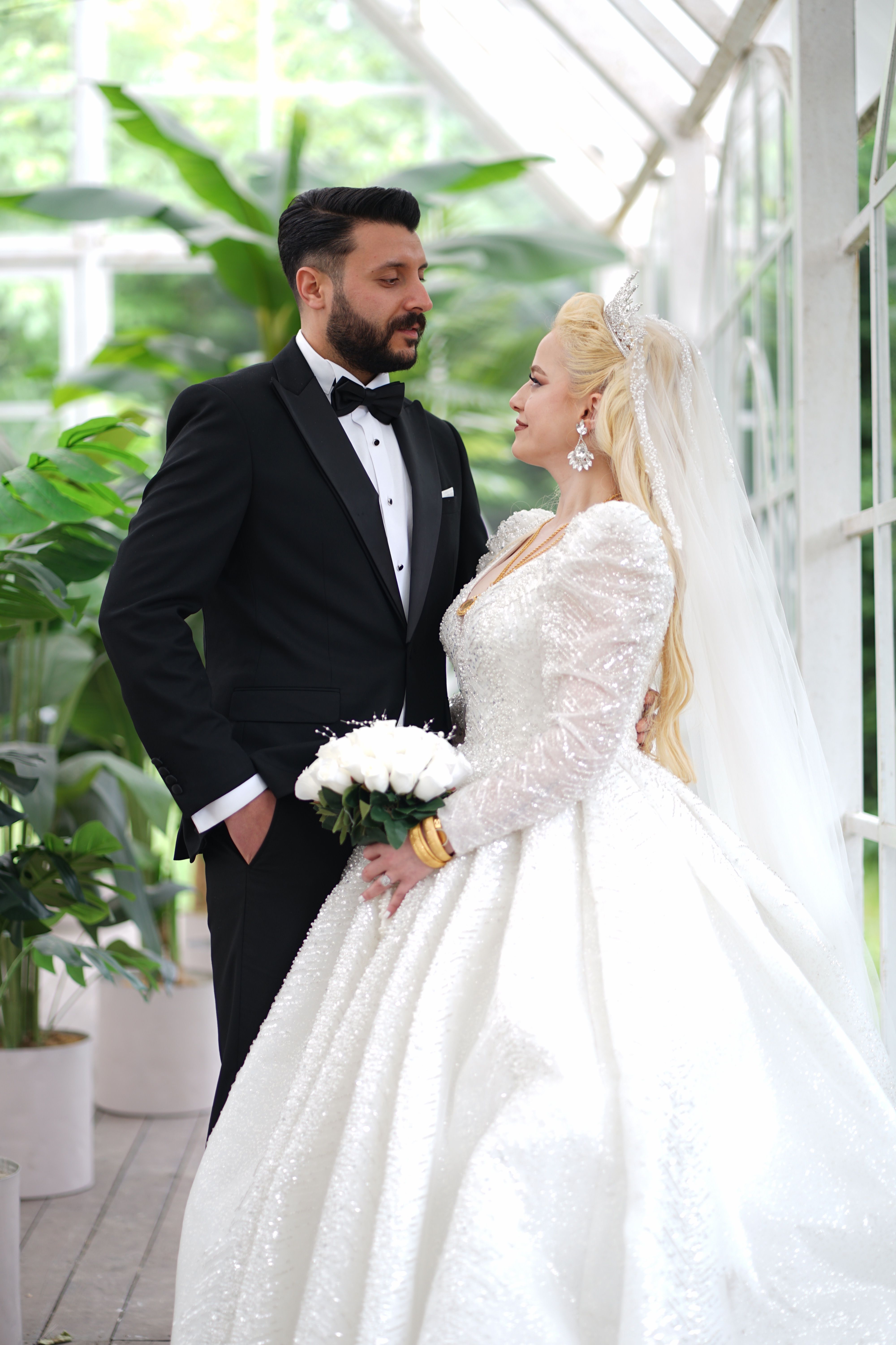 A bride and groom elegantly pose in a lush, indoor setting, exuding romance and style.
