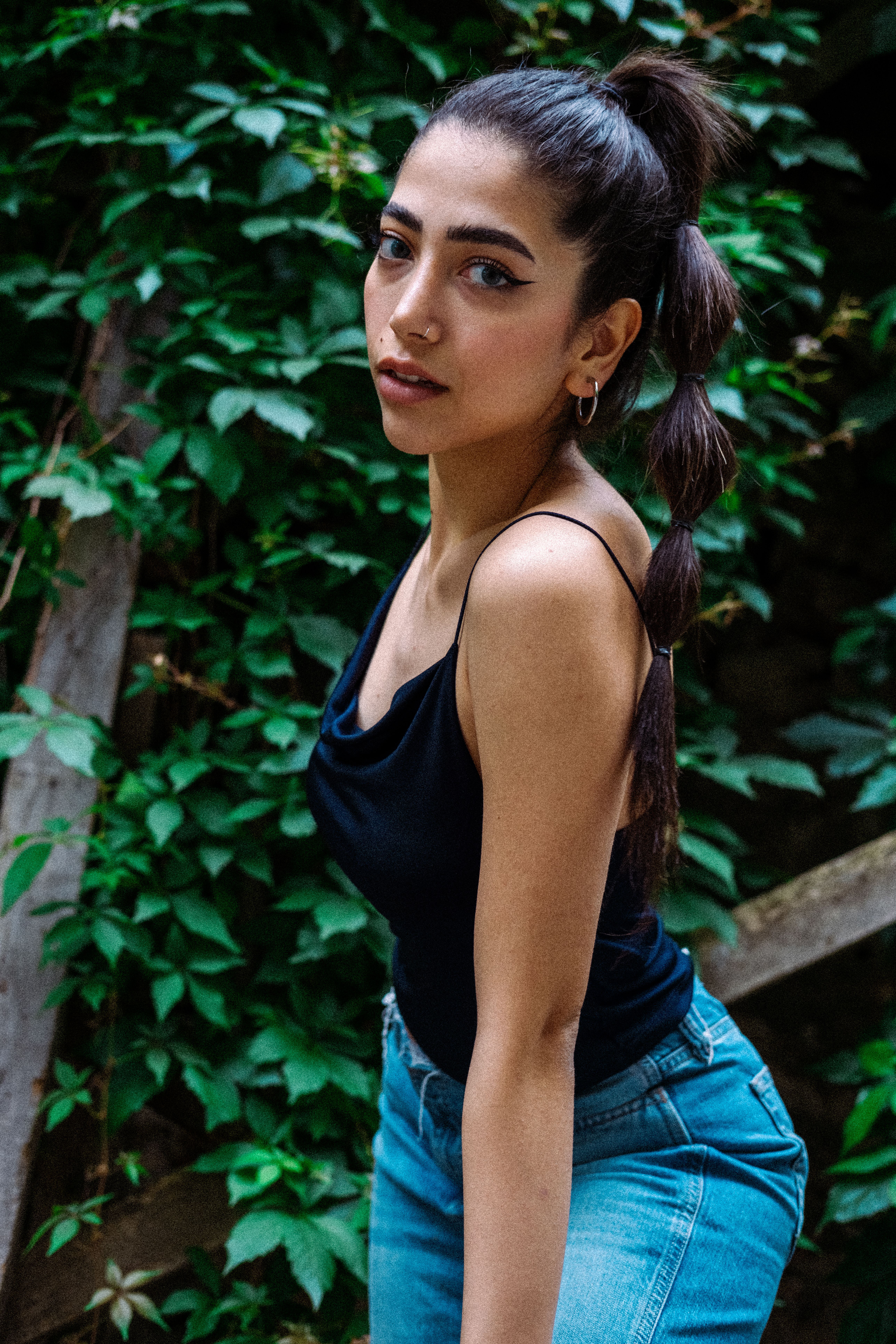 Elegant woman in black top and jeans posing against lush green leaves outdoors.