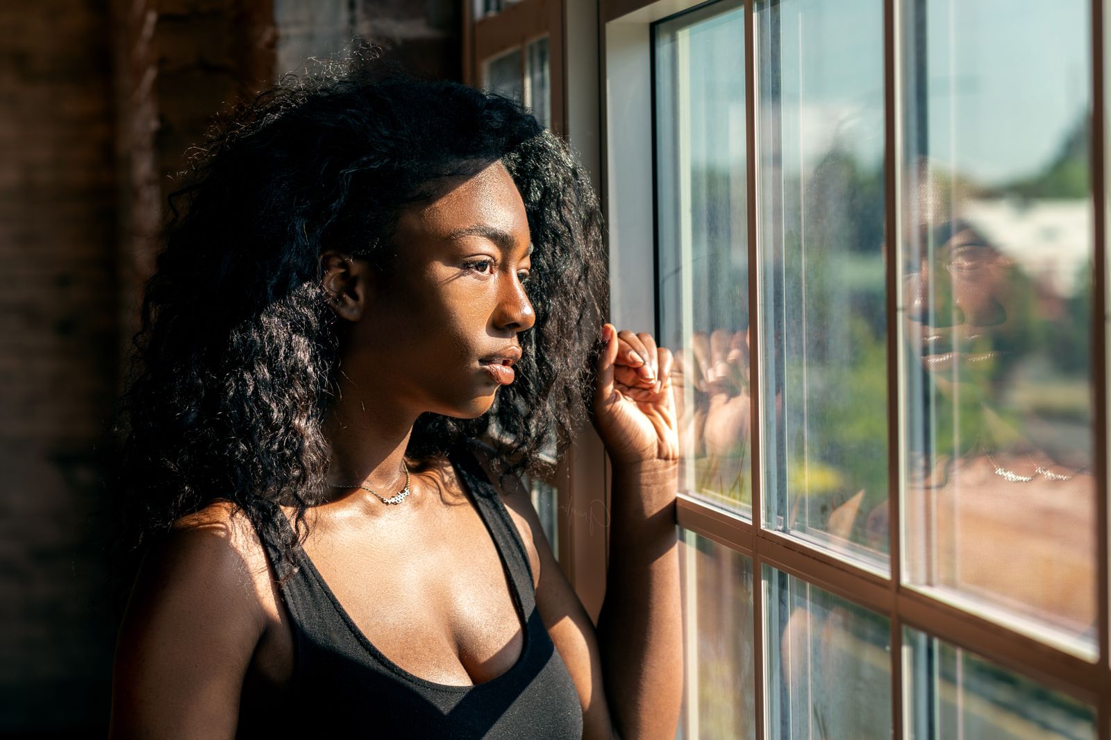 Woman with curly dark hair looking thoughtfully out a sunlit window with warm afternoon light.