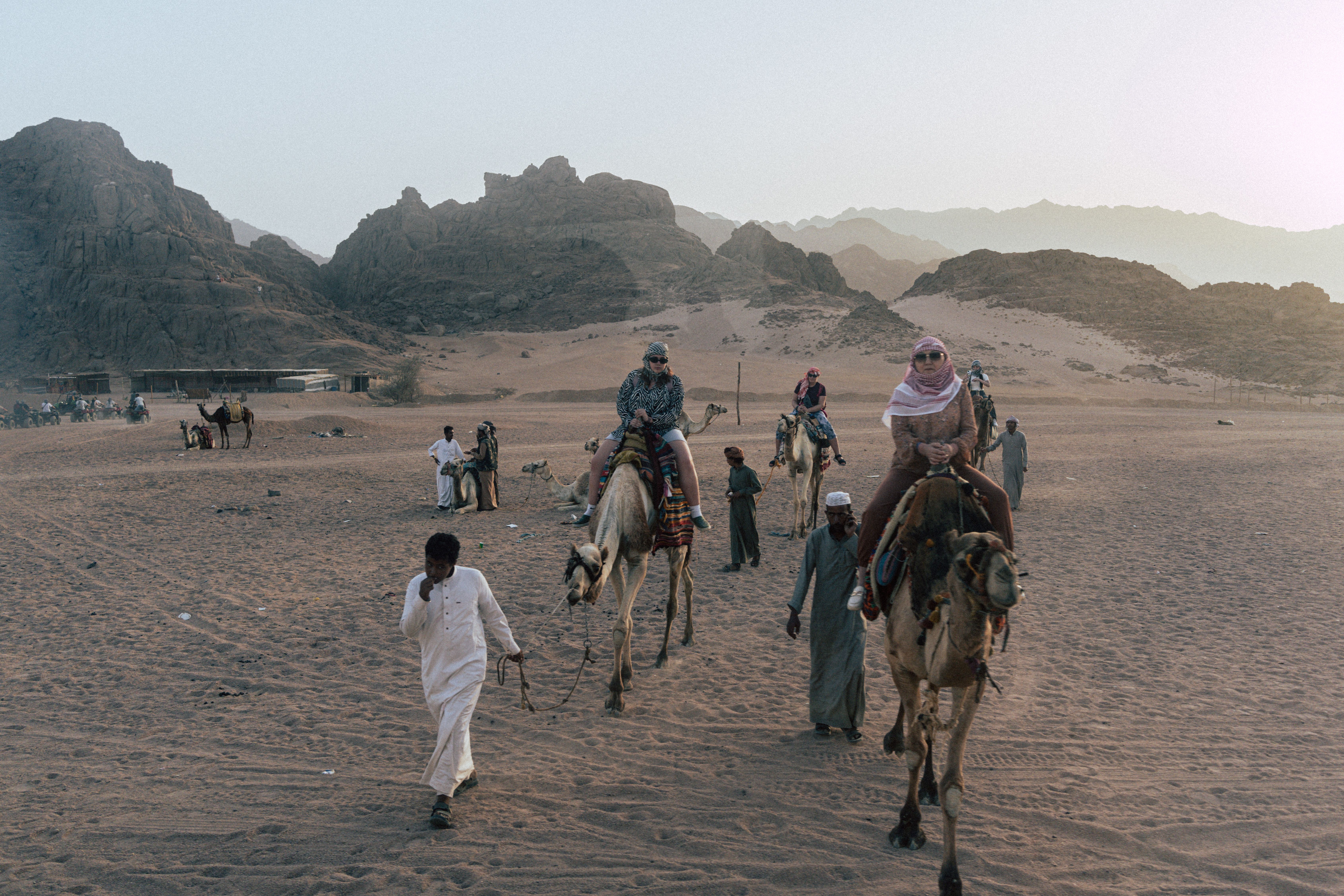 Adventure seekers ride camels through a sandy desert landscape with rocky mountains in the backgroun
