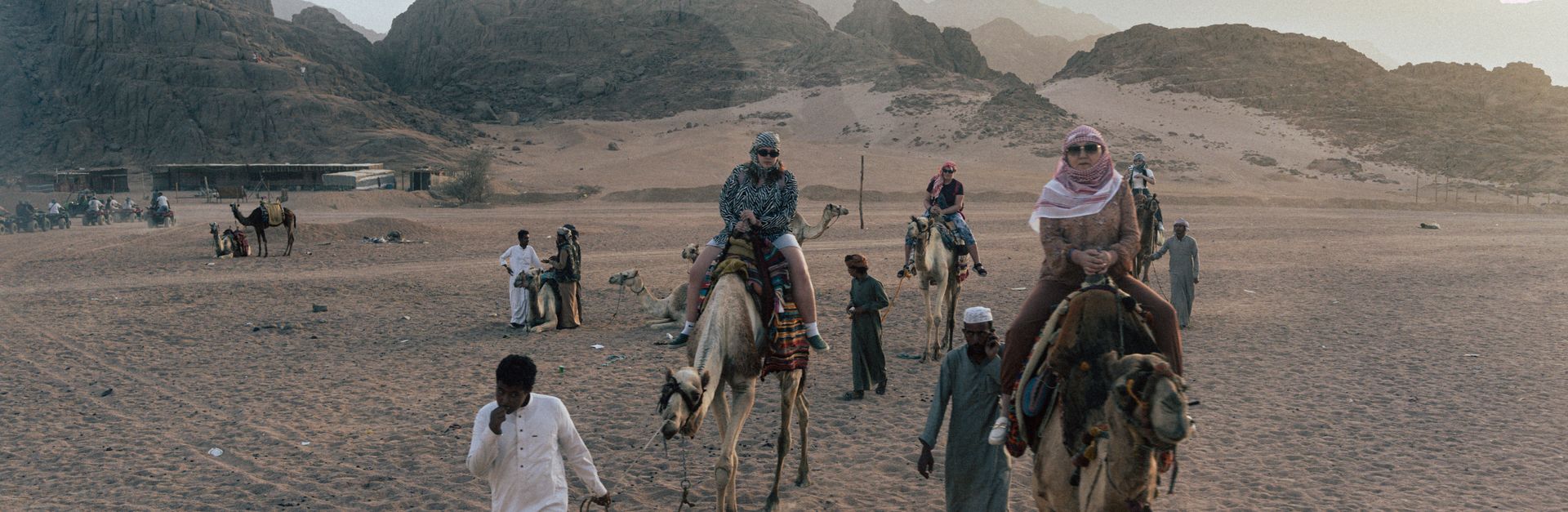 Adventure seekers ride camels through a sandy desert landscape with rocky mountains in the backgroun