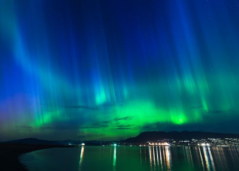 Vibrant aurora borealis illuminating the night sky above a city by the sea.