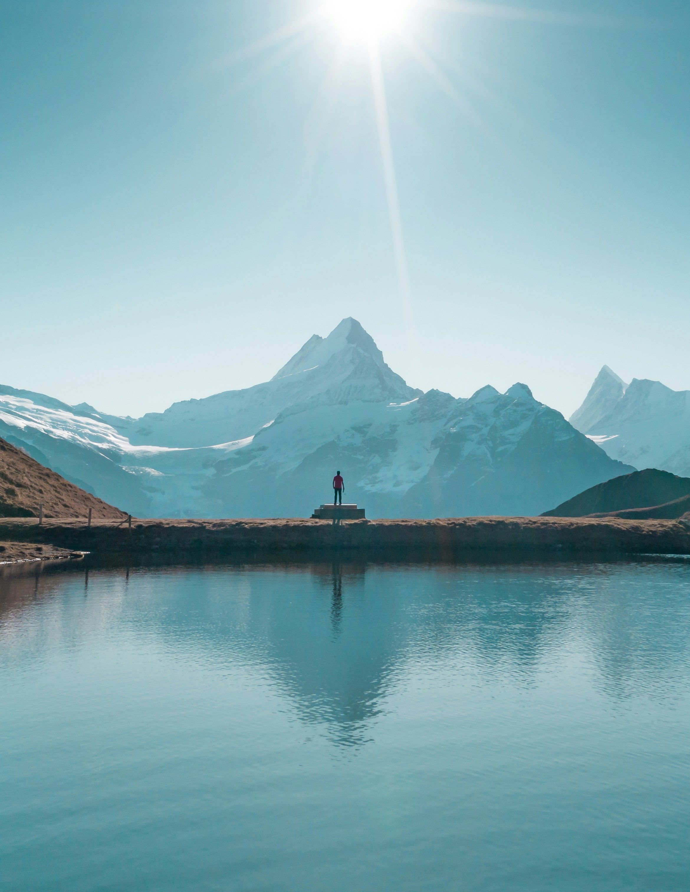 A lone hiker stands by a tranquil lake, facing stunning snowcapped mountains under a bright sky.