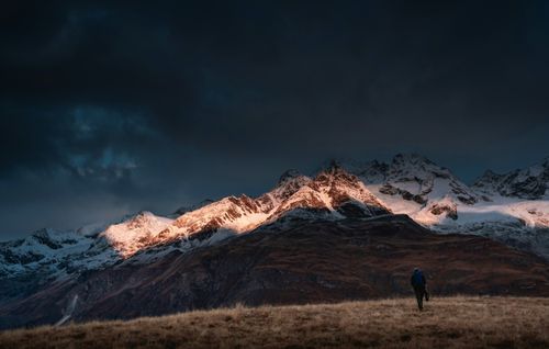 Free A lone hiker walks toward stunning snow-capped peaks at sunset, under dramatic skies. Stock Photo