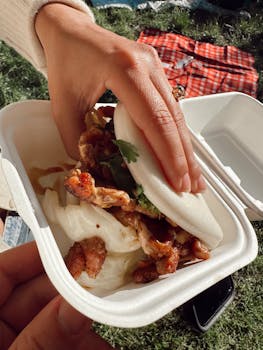 Close-up of a bao bun with fried chicken and cilantro in an outdoor picnic setting.