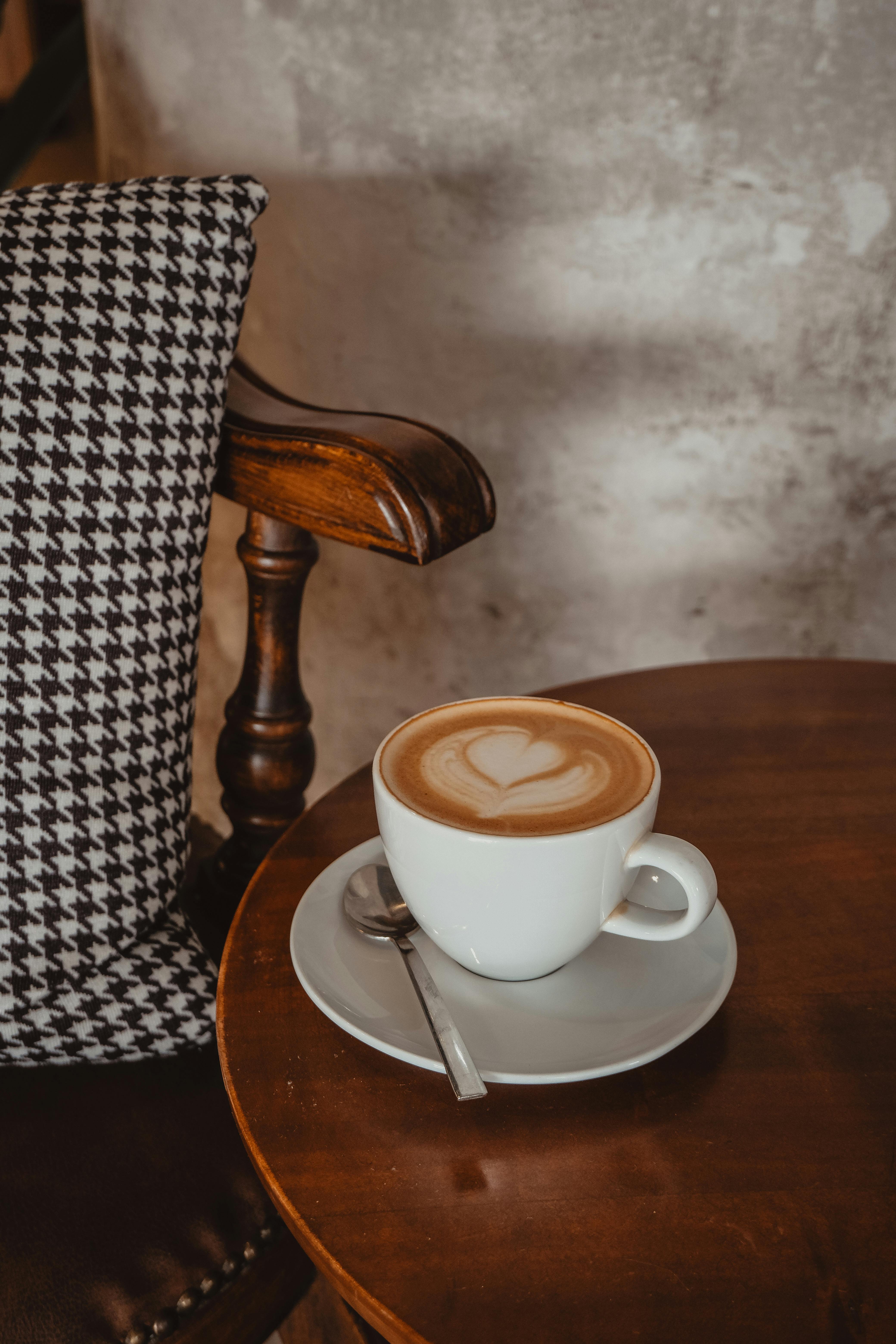 A warm cup of cappuccino with heart froth art on a wooden table, next to a vintage chair.