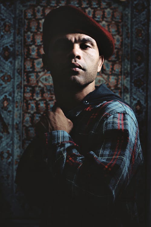 Serious bearded ethnic male in checkered shirt and red beret standing in dark place and looking at camera confidently