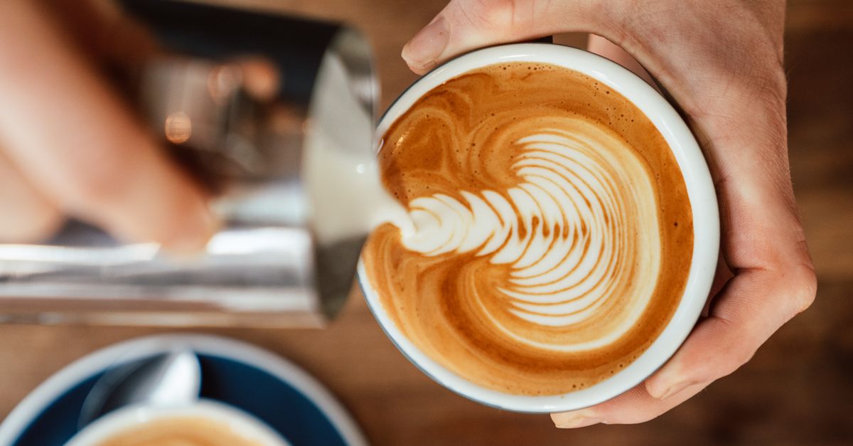 Close-up of barista pouring milk creating intricate latte art in coffee cup.
