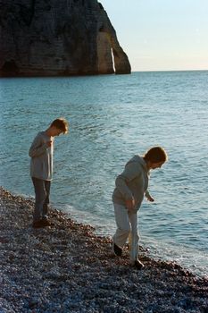 Two children walking along the pebbled shore near iconic cliffs at Étretat, Normandy, France.