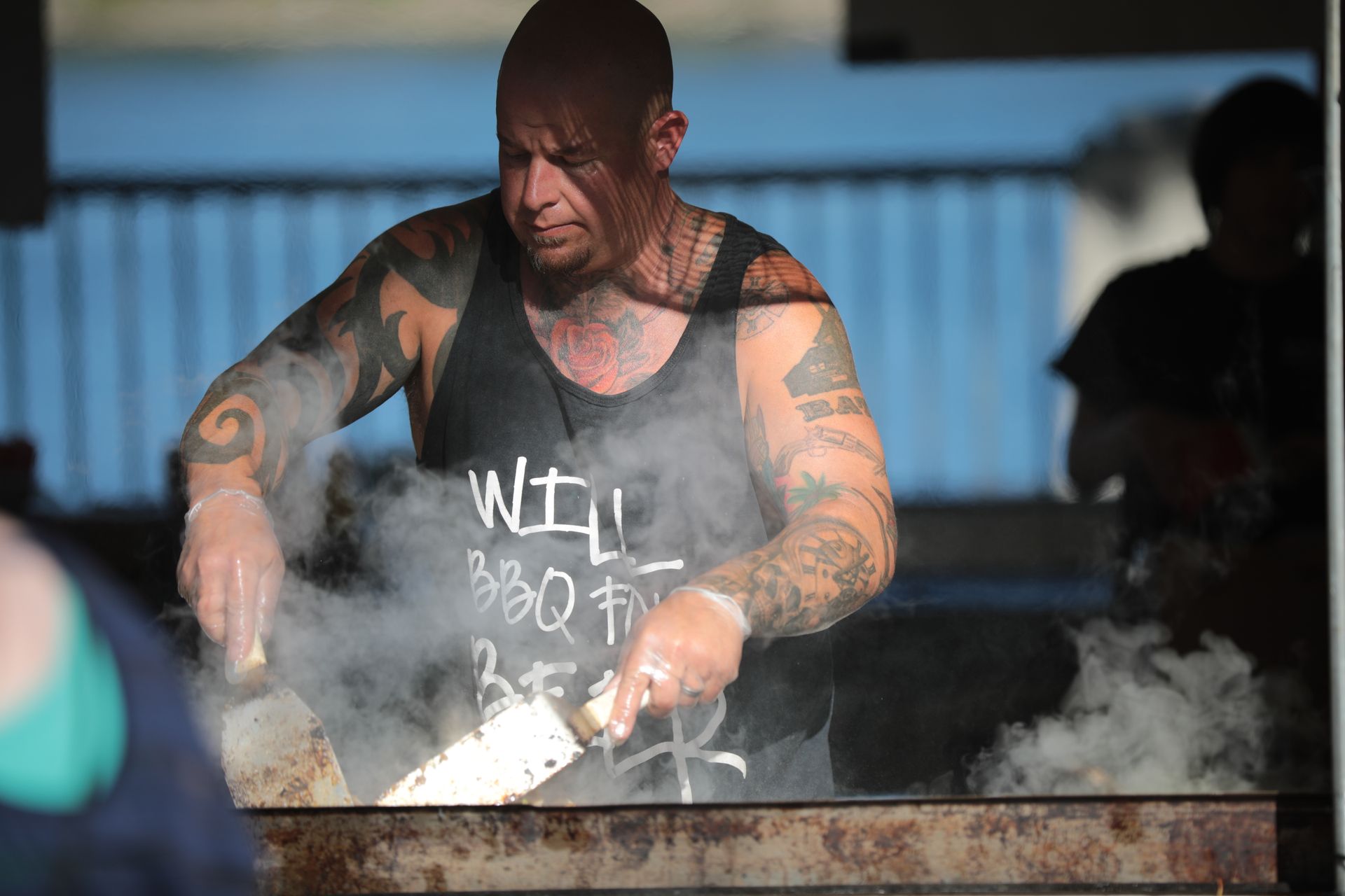 A tattooed chef grills food outdoors, surrounded by smoke and natural light.