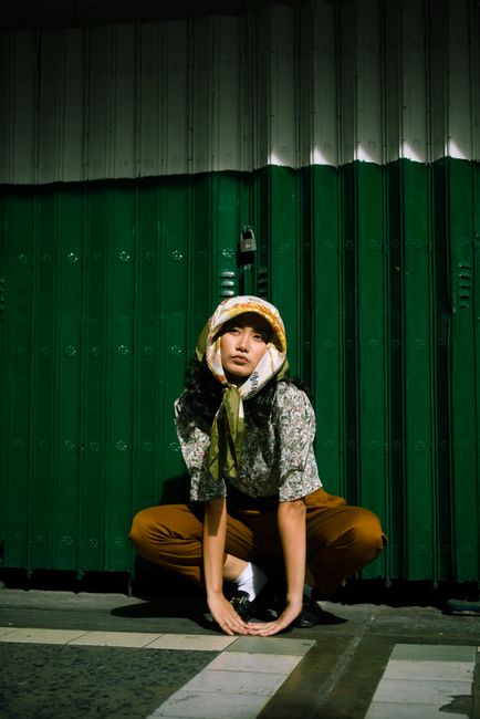 Fashionable woman crouching outdoors against green steel shutters, wearing a floral blouse and bandana.