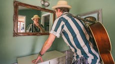 室内，一位戴着软毡帽的音乐家若有所思地站在镜子旁，手里拿着一把原声吉他