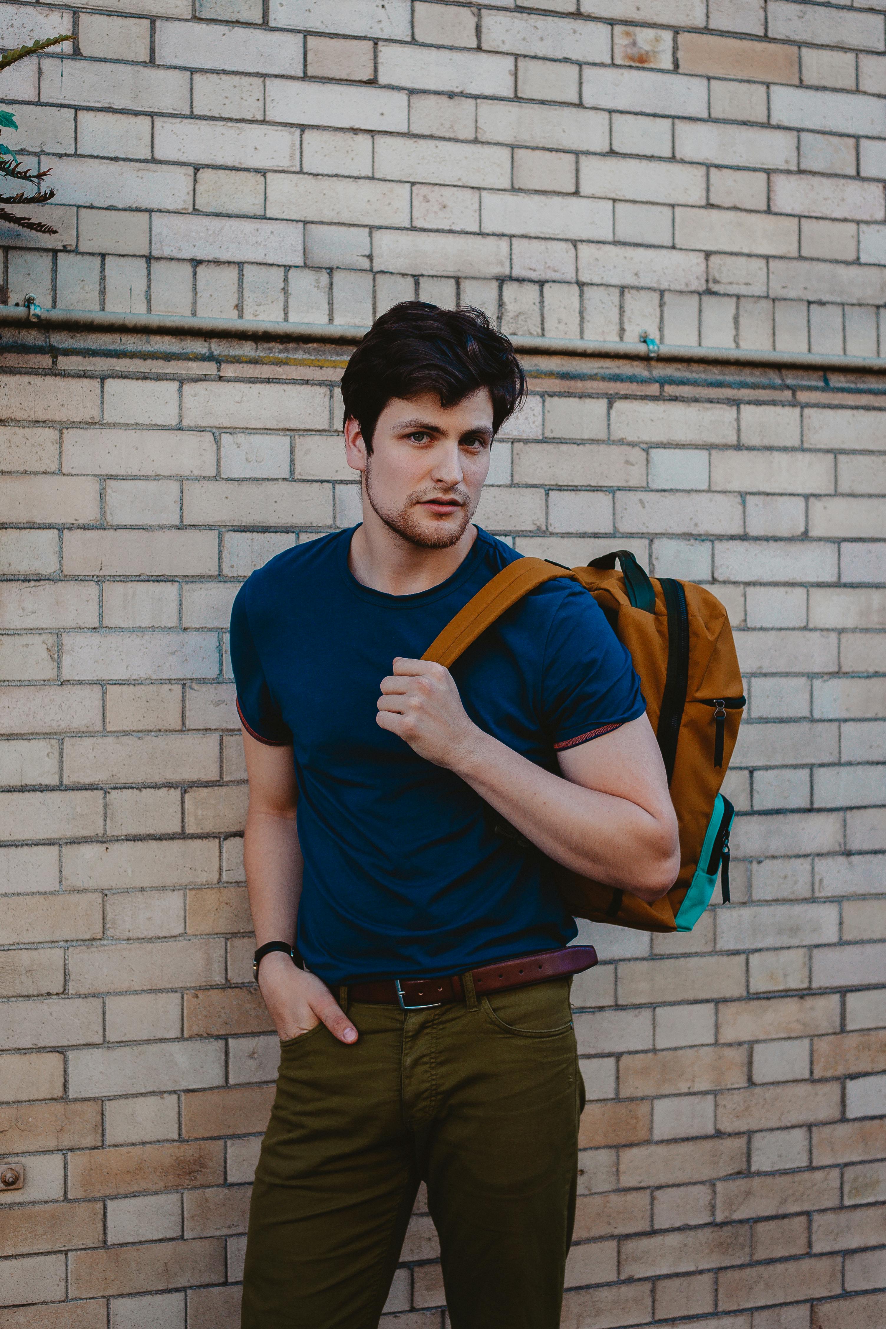 一名穿着蓝色衬衫、背着背包的男子随意地靠在砖墙上
