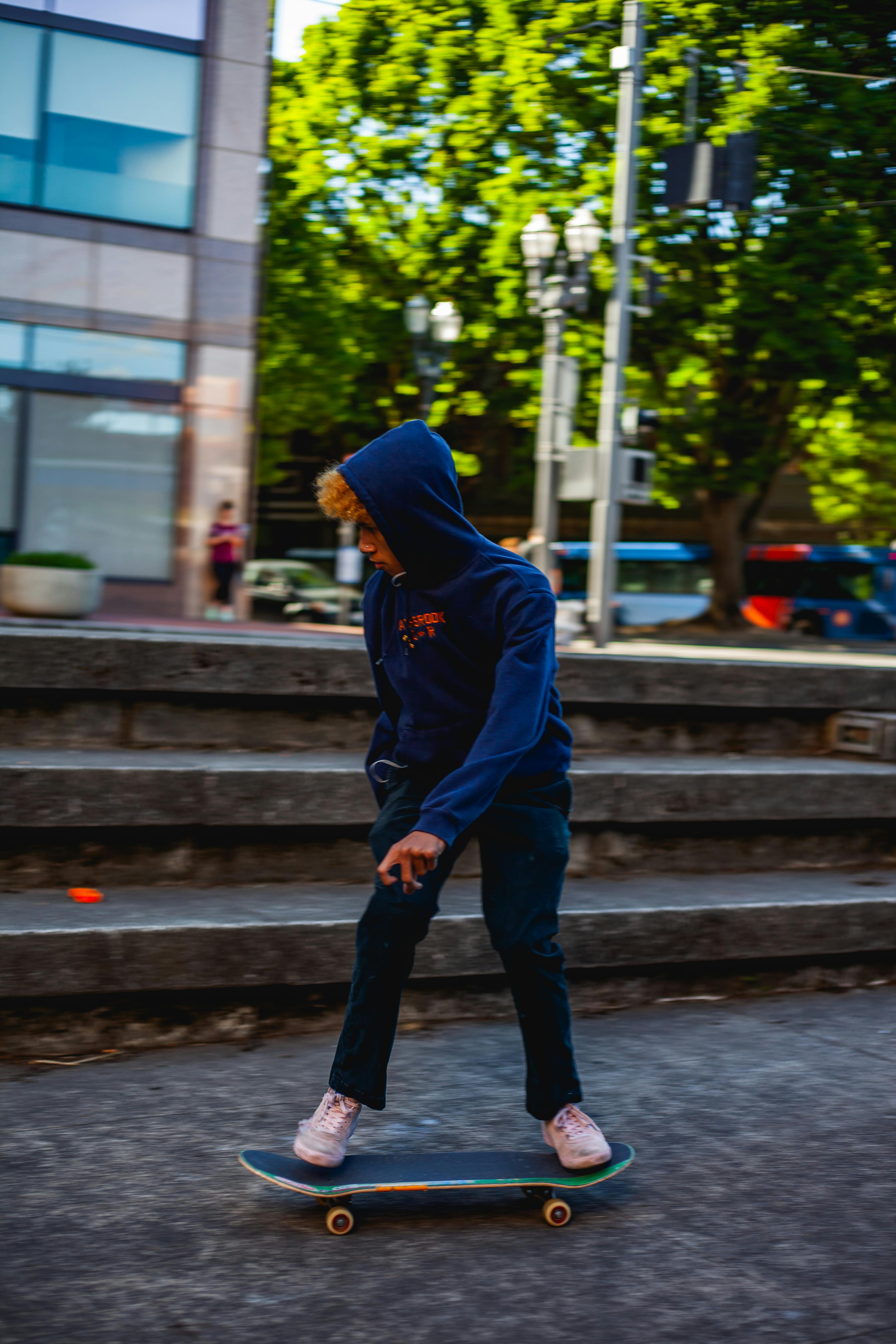 Young man skateboarding on city streets with vibrant background trees and urban architecture.