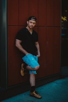A stylish man posing outdoors in a casual outfit with a baseball cap and shorts.