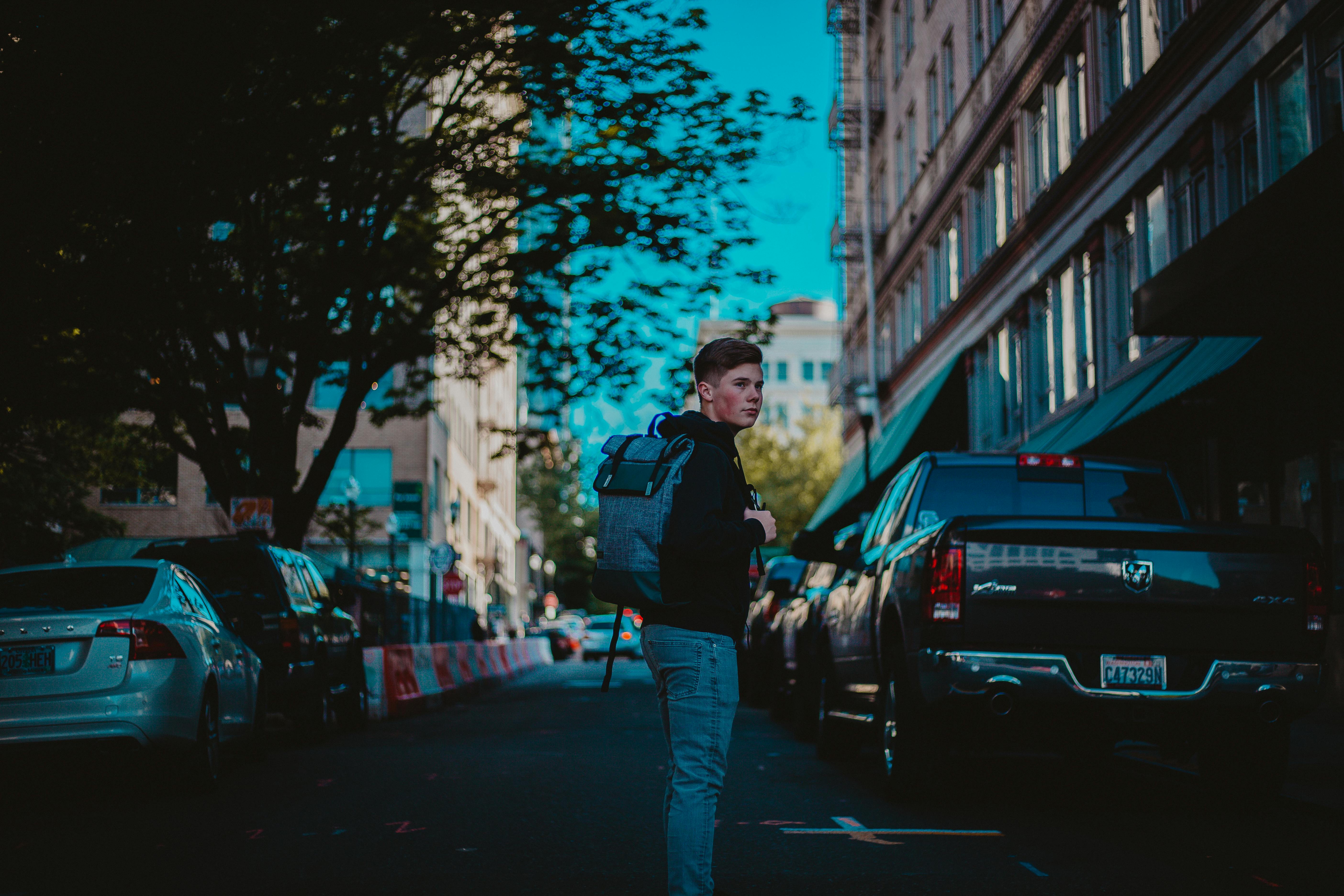 A young man carrying a backpack walks in an urban street, surrounded by parked cars.