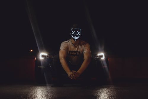 Free A man wearing an illuminated LED mask sits in front of a car with headlights on at night. Stock Photo