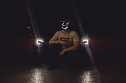 Free A person wearing an LED mask poses by a car with illuminated headlights at night, creating a spooky vibe. Stock Photo