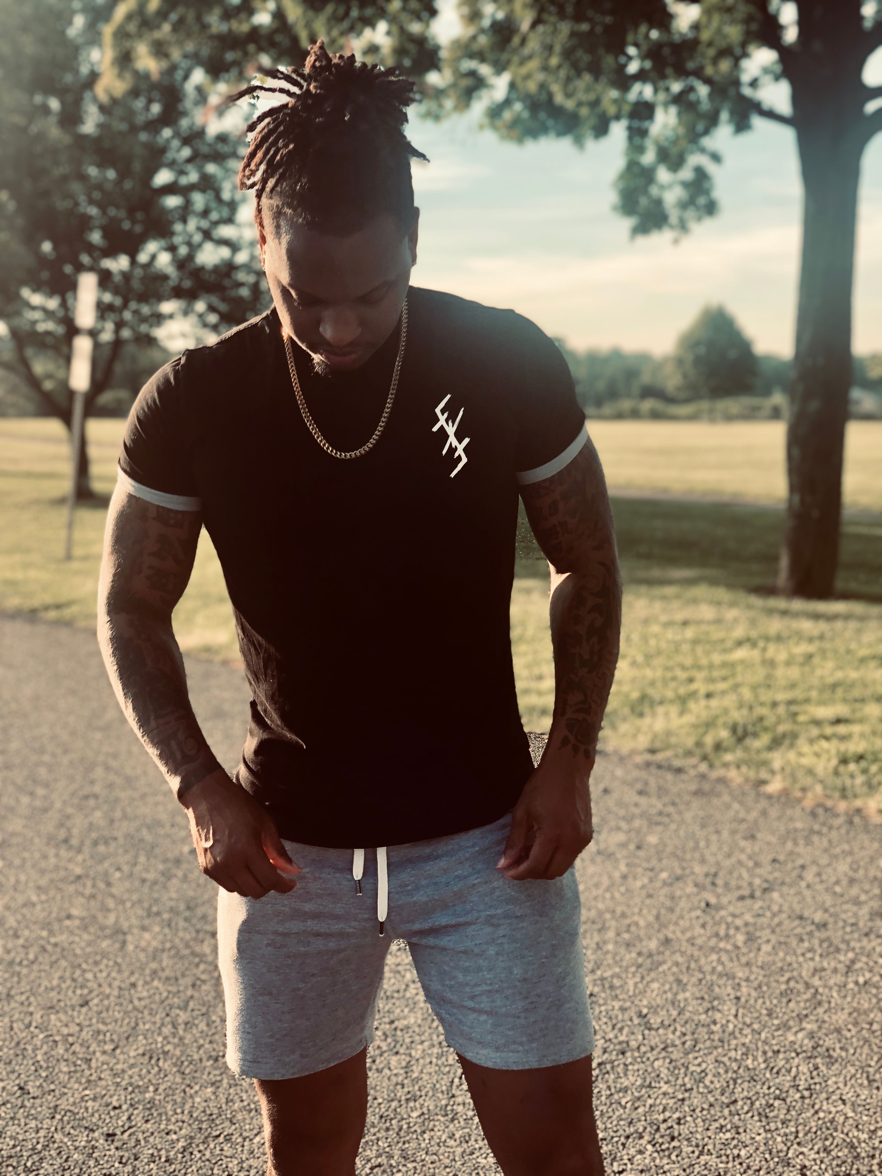 Fashionable tattooed man in casual attire stands outdoors during the day, showing style and confidence.