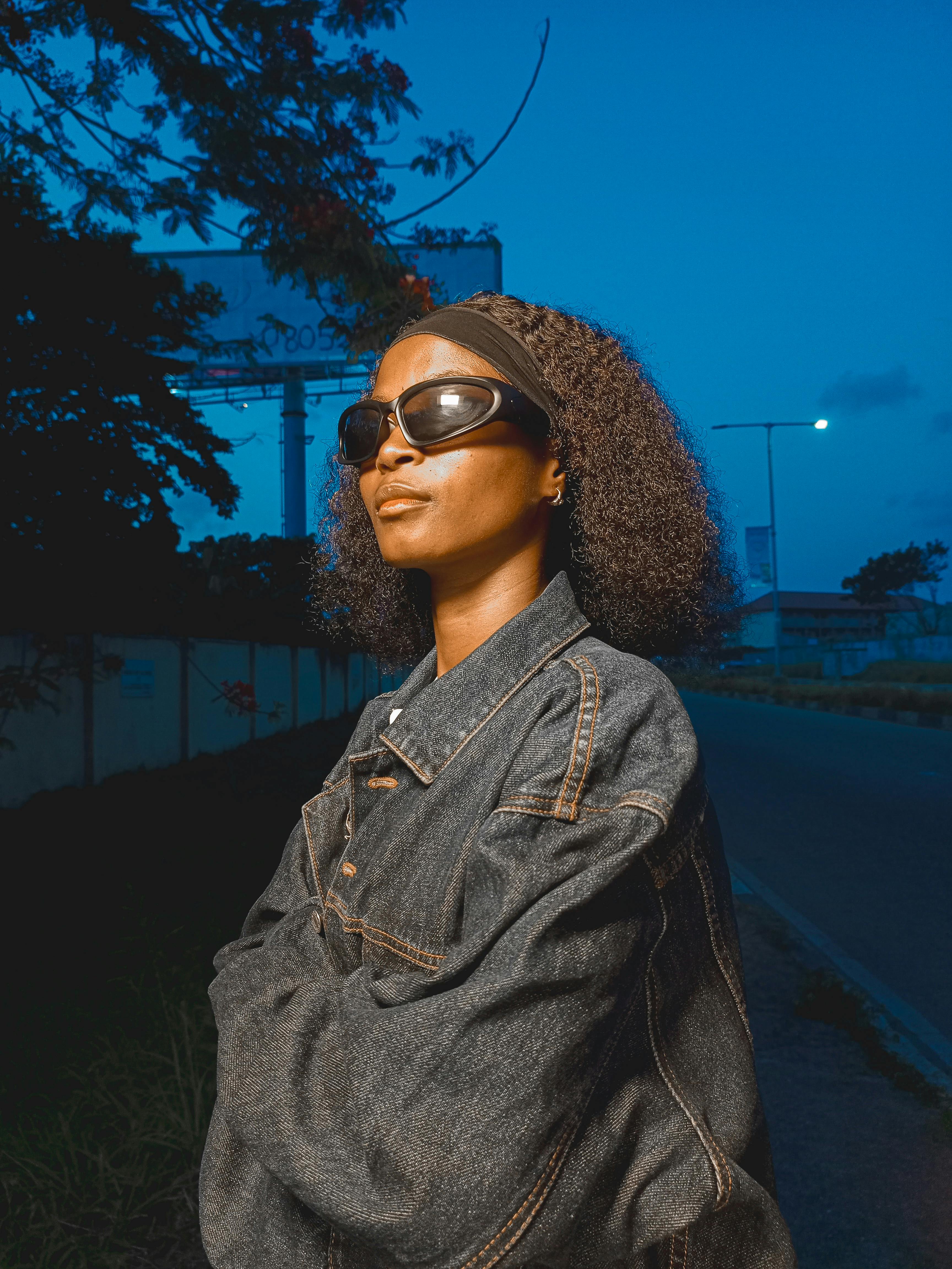 Free Confident woman in sunglasses and denim jacket posing outdoors during twilight, showcasing urban fashion. Stock Photo