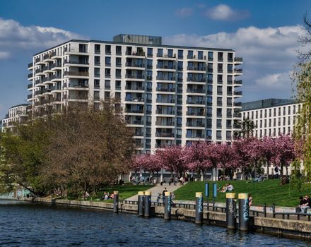 A contemporary apartment building by the river in Berlin, surrounded by spring blossoms and lively park ambiance.