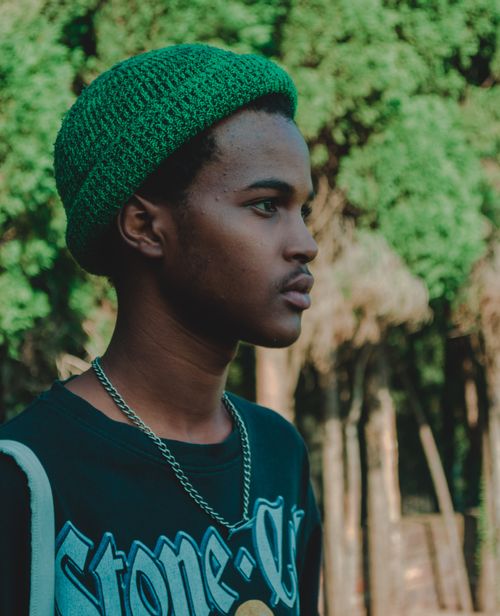 Free Profile view of a stylish young man wearing a green knit cap and necklace, standing outdoors in nature. Stock Photo