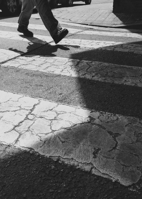 Black and white image of a person walking on a pedestrian crossing, casting a shadow on the cracked pavement.