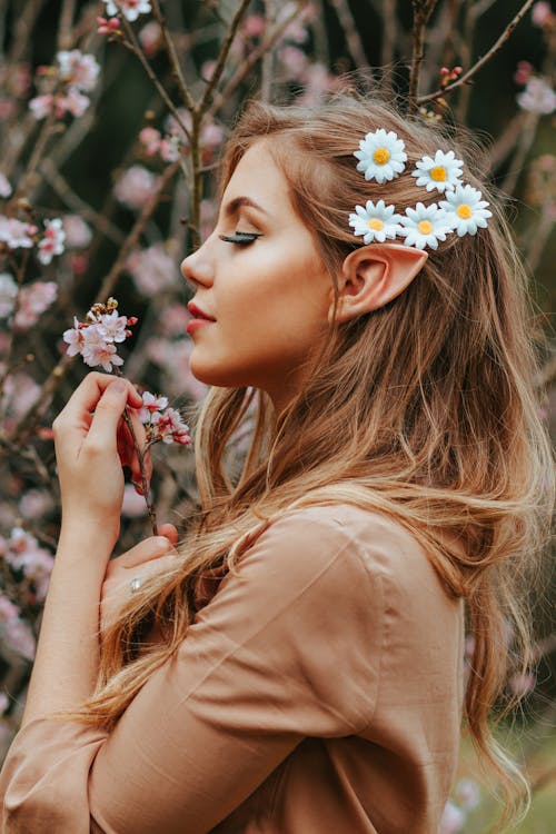 Side View Photo of Woman with Pointy Ear and Daisies on Her Hair Standing by Cherry Blossoms While Holding Cherry Blossom Branch