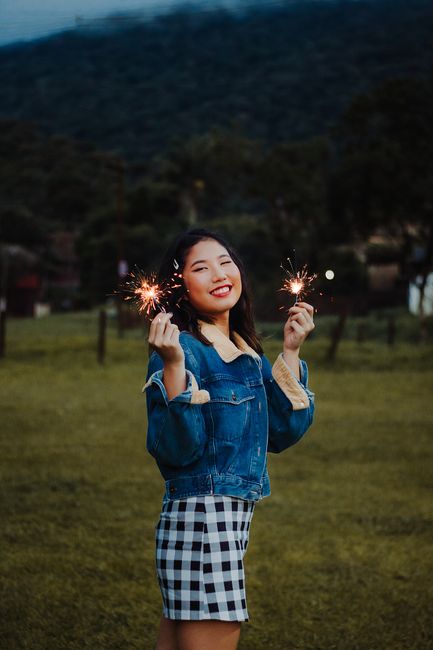 Smiling Asian woman in a checkered skirt and denim jacket holding sparklers in a scenic outdoor setting.